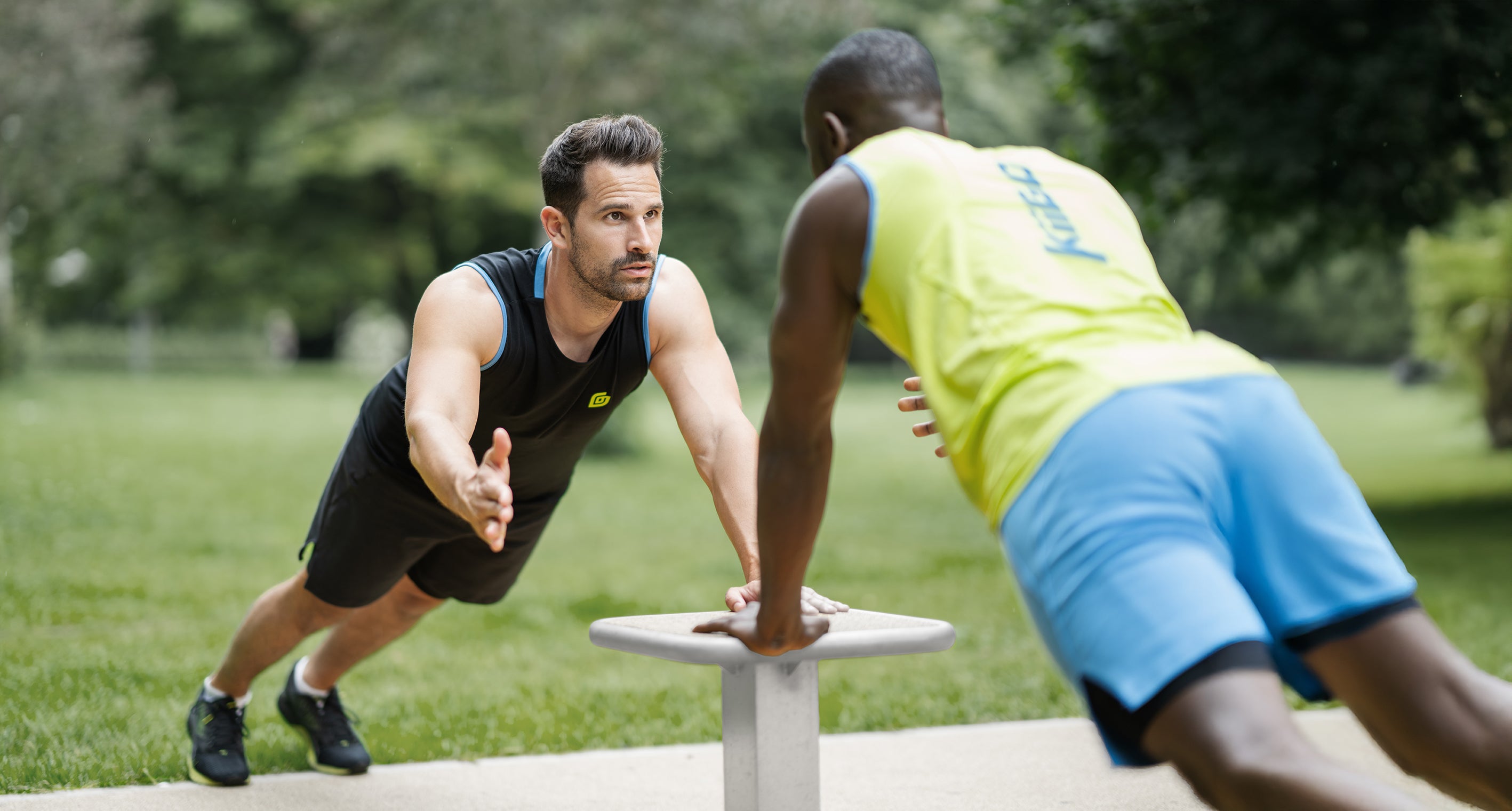 2 Sportler im Calisthenics Park. Sie klatschen sich ab, während sie an einem Gerät Push ups machen. Ein Sportler trägt das schwarze KiiGO Herren Tanktop mit der schwarzen KiiGO 2-in-1 Short, der andere Sportler trägt die KiiGO Kombination mit KiiGO Tanktop in Lime und KiiGO Short 2-in-1 in blau.