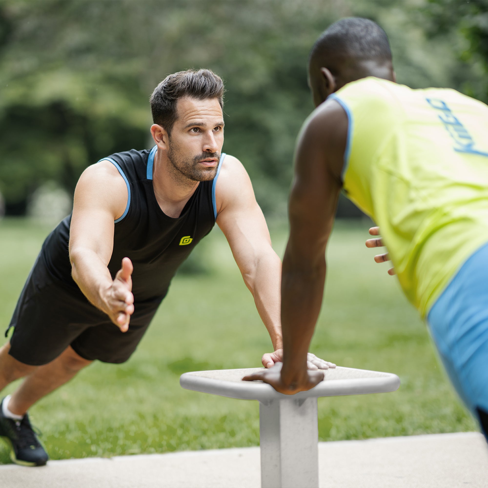 Zwei Männer beim intensiven Outdoor-Workout im Park: Der Fokus liegt auf dem Athleten im nachhaltigen und schwarzen KiiGO Herren Sport Tanktop beim Liegestütz machen. Im Vordergrund ist das neongrüne Tanktop (Limepunch grün) in der Rückenansicht zu sehen. Nachhaltige Activewear aus Tencel.