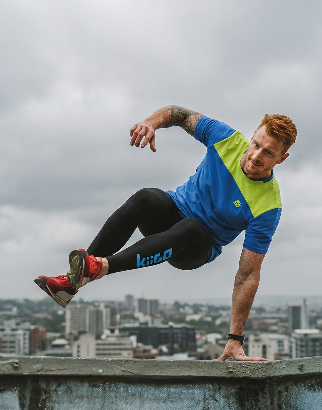 Ein junger Mann springt beim Laufen über eine Mauer. Er trägt dabei das KiiGO Herren Loose-Tee in blau und die KiiGO Herren Leggins in Schwarz.
