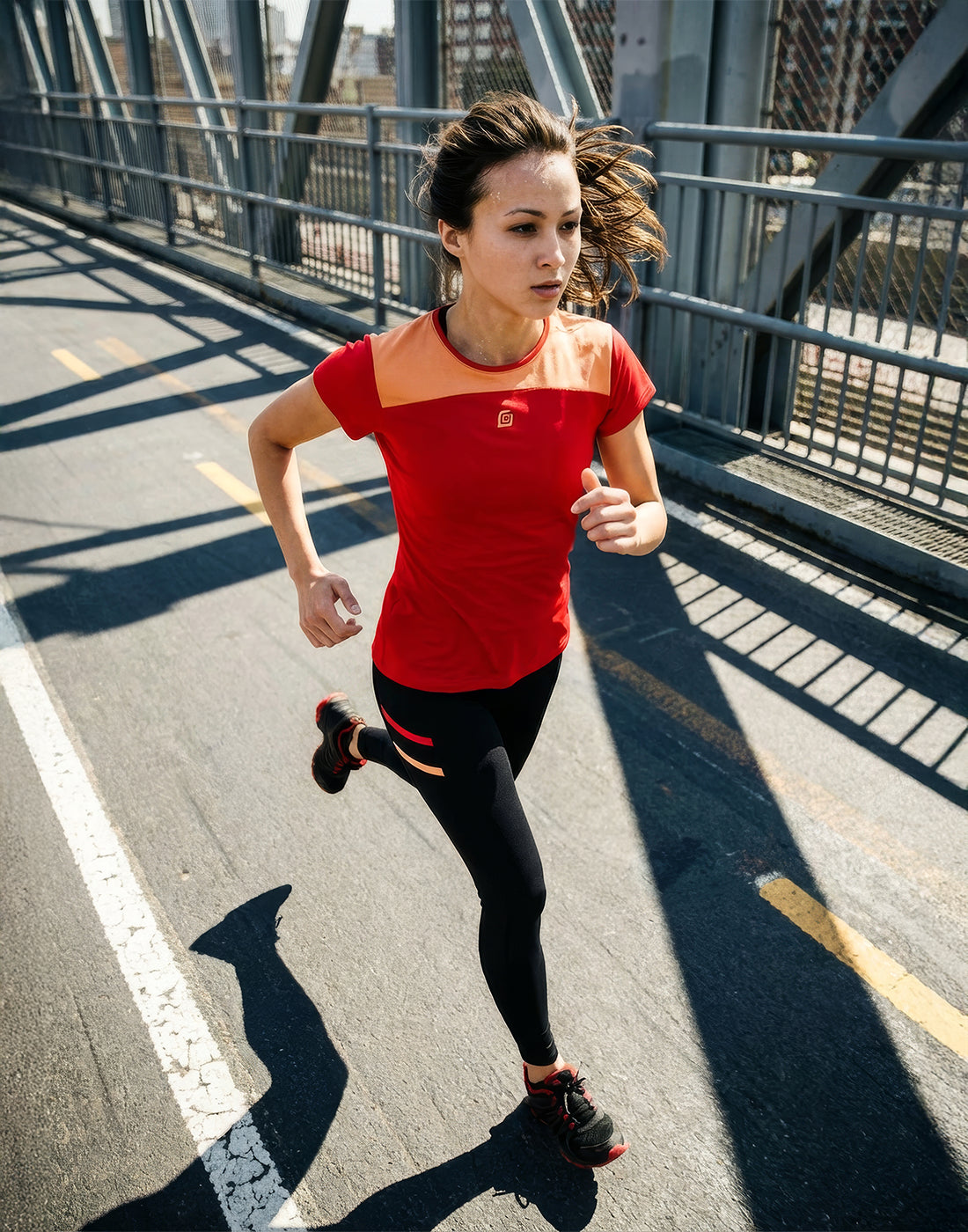 Läuferin joggt über eine Brücke. Sie trägt das rote KiiGO Damen Shirt und die passende KiiGO Damen Leggins dazu. Die Sonne scheint.
